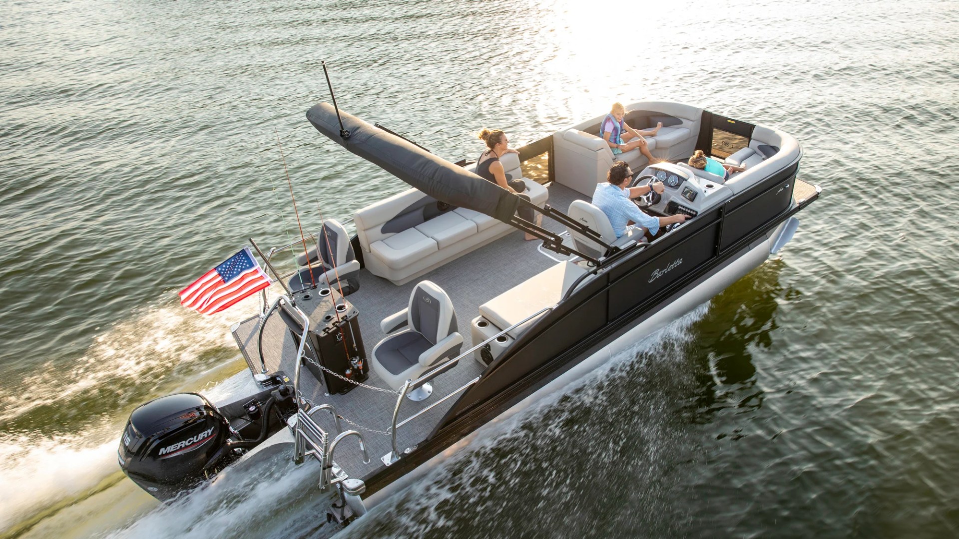 Pontoon boat with four people cruising on a lake, American flag flying at the back.