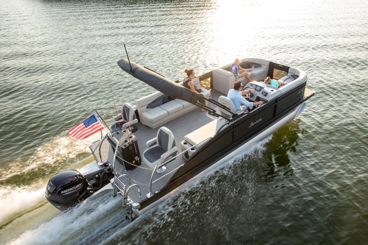 Pontoon boat with four people cruising on a lake, American flag flying at the back.
