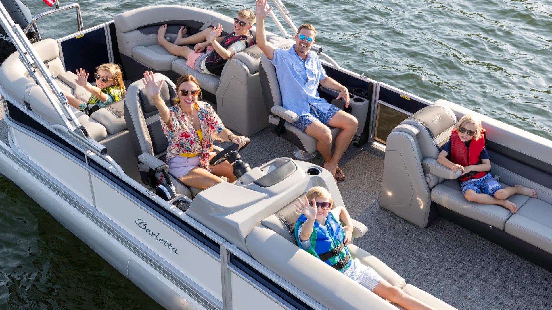 Family of six waves while seated on a pontoon boat on a sunny day.
