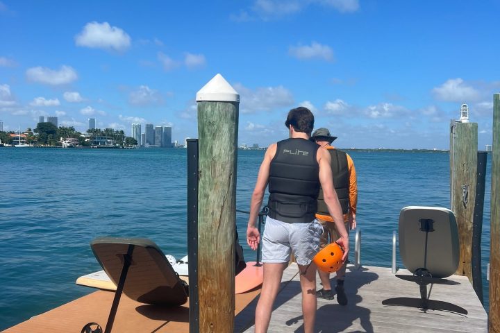Two people on a dock with paddleboards and bright blue sky.