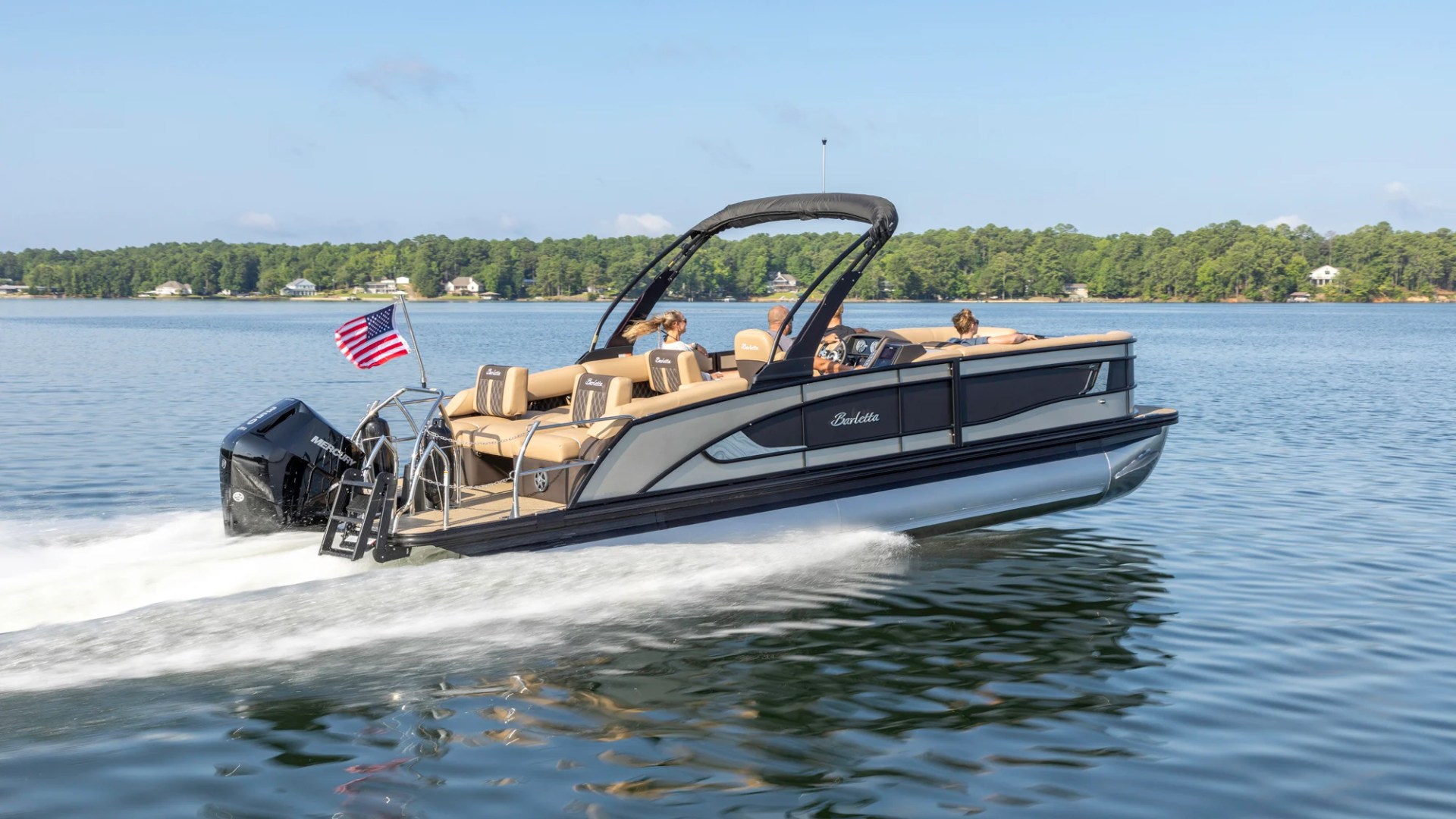Pontoon boat with people cruising on a lake, American flag flying at the rear.