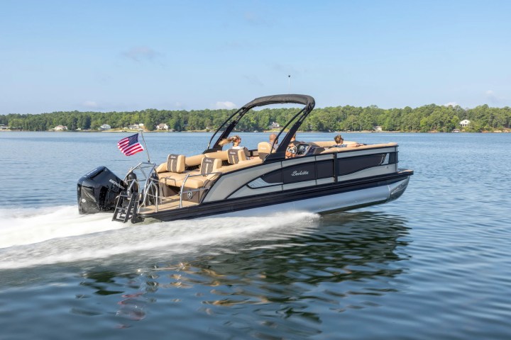 Pontoon boat with people cruising on a lake, American flag flying at the rear.