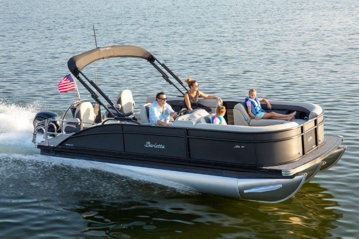 Family rides a pontoon boat with an American flag on a sunny lake.