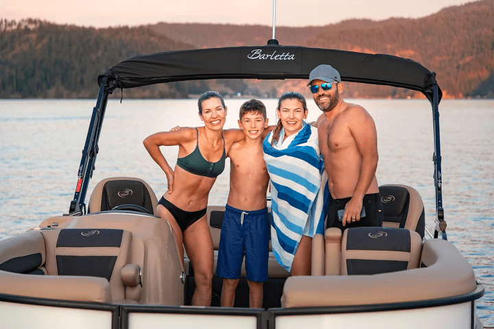 Four people in swimsuits on a boat, with a scenic lake and mountains in the background.