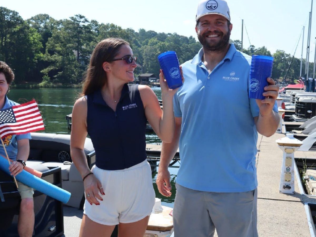 People smiling on a dock holding blue cups, one with a cap, yacht in background.